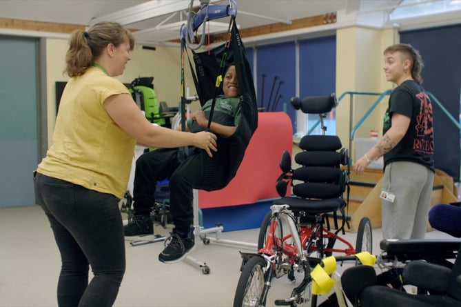 Student Caleb uses a hoist to move on to a trike, supported by head of physiotherapy Clare Bradford and physiotherapy assistant Amy Trussler, Treloar's, Holybourne, December 2024.